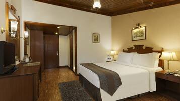 Habitación de hotel con techo de madera, cama con ropa blanca, mesita de noche con lámpara, escritorio con ordenador y una alfombra pequeña.