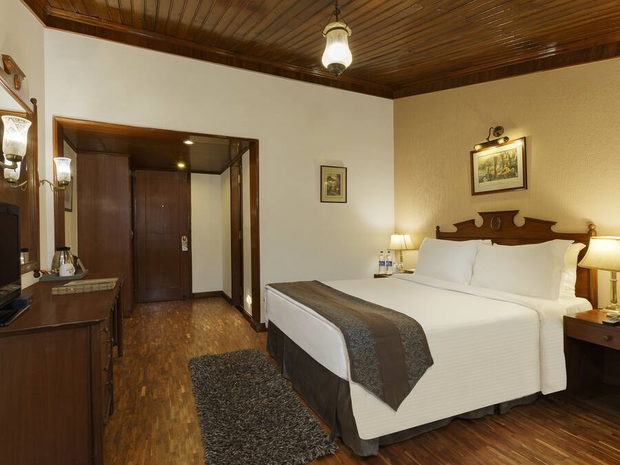 Ein Hotelzimmer mit Holzboden, einem Bett mit weißen Leinen, einem Nachttisch mit Lampe, einem Schreibtisch mit Computer und einem kleinen Teppich.