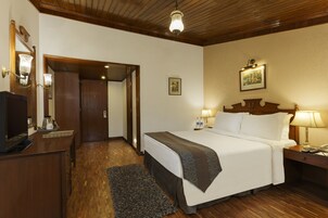 Habitación de hotel con techo de madera, cama con ropa blanca, mesita de noche con lámpara, escritorio con ordenador y una alfombra pequeña.
