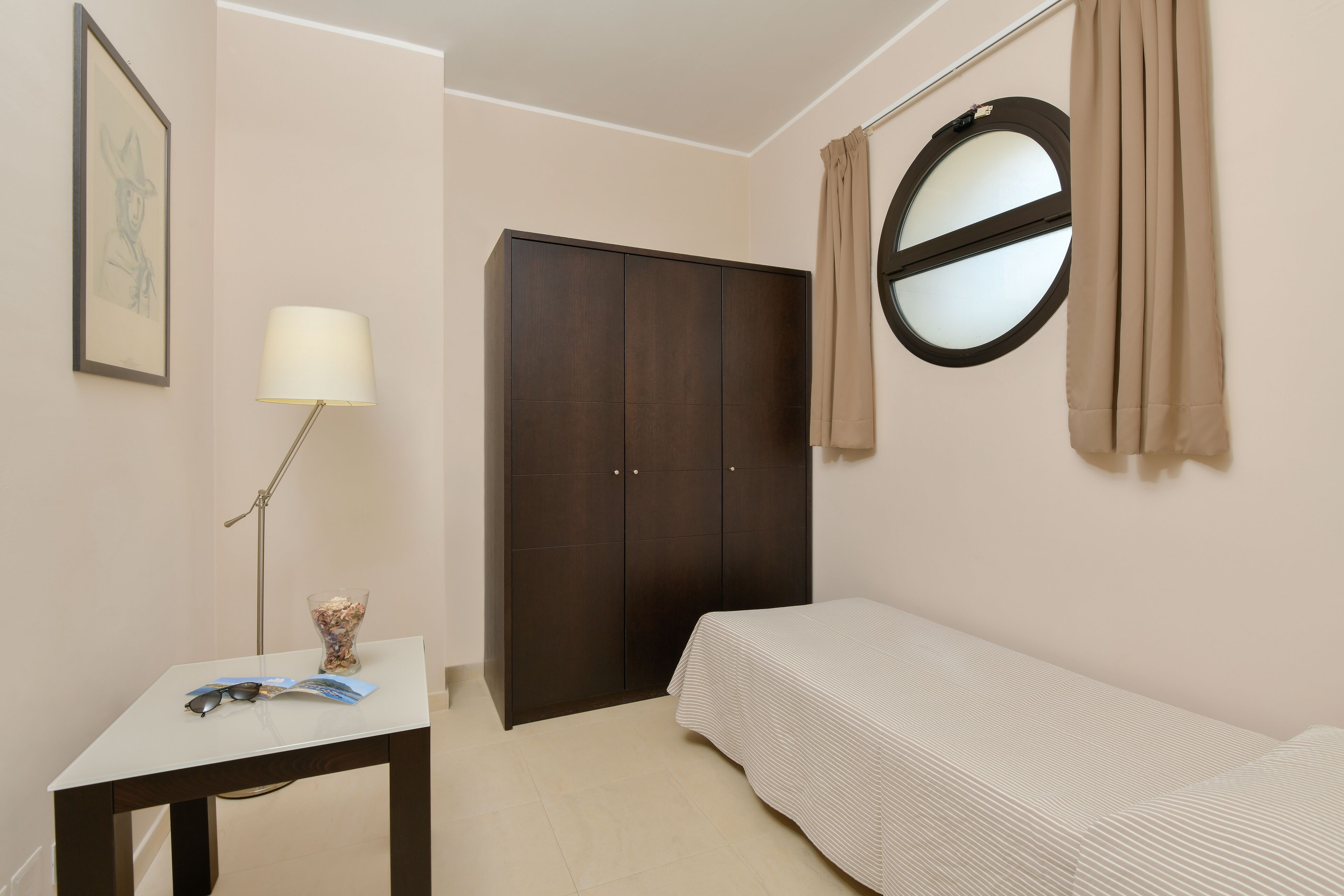 A small, neatly arranged bedroom with a single bed, a dark wardrobe, a bedside table with a lamp, and a framed picture on the wall.