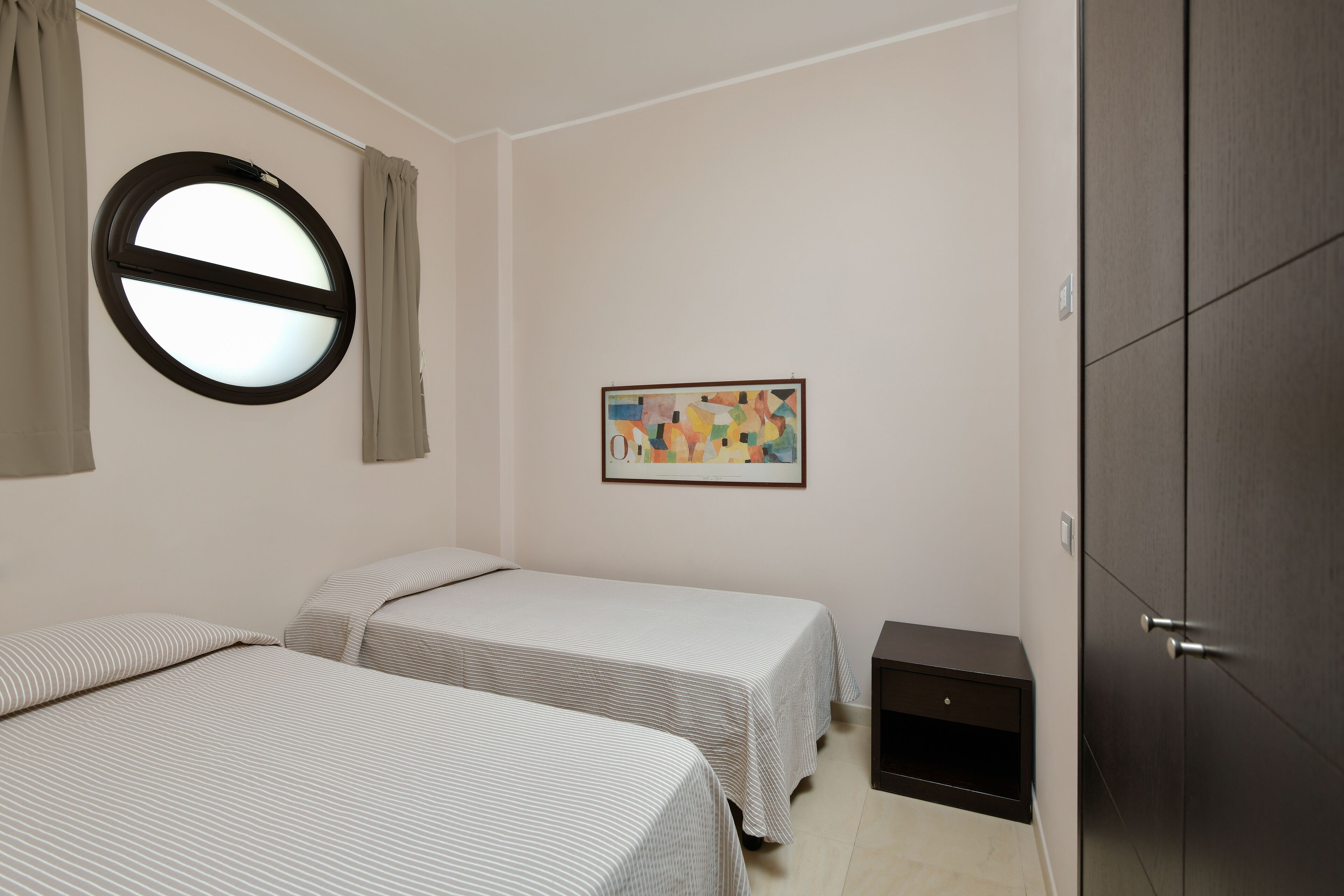 Une chambre avec deux lits, une fenêtre ronde, un tableau et une armoire.