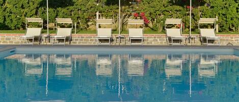 A poolside area with lounge chairs and umbrellas, a stone wall, and a hedge with red flowers.