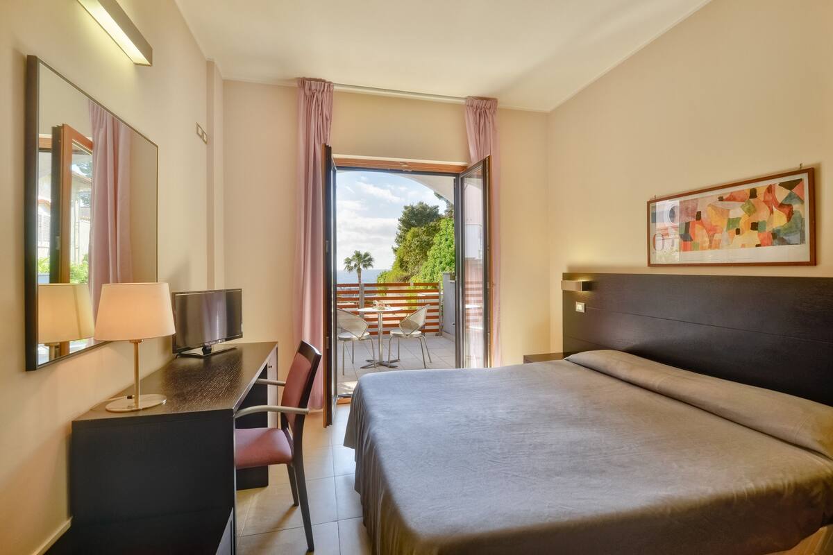A hotel room with a bed, desk, chair, and a balcony with a view of greenery.