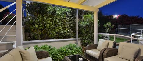 Une terrasse extérieure aménagée avec des meubles en osier, une table basse en verre et une vue sur un jardin au crépuscule.