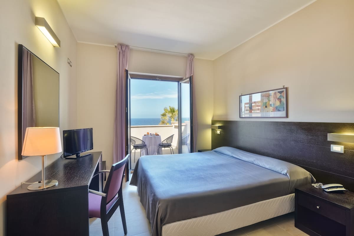 A hotel room with a bed, a desk with a lamp, a TV, a chair, a small table, and a balcony with a view of the sea.