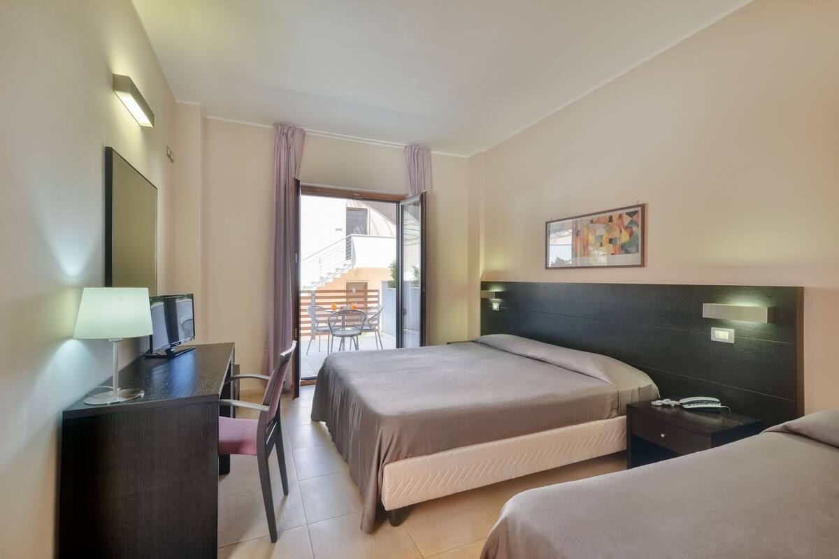 A hotel room with two beds, a desk with a lamp, a TV, and a balcony with a view.