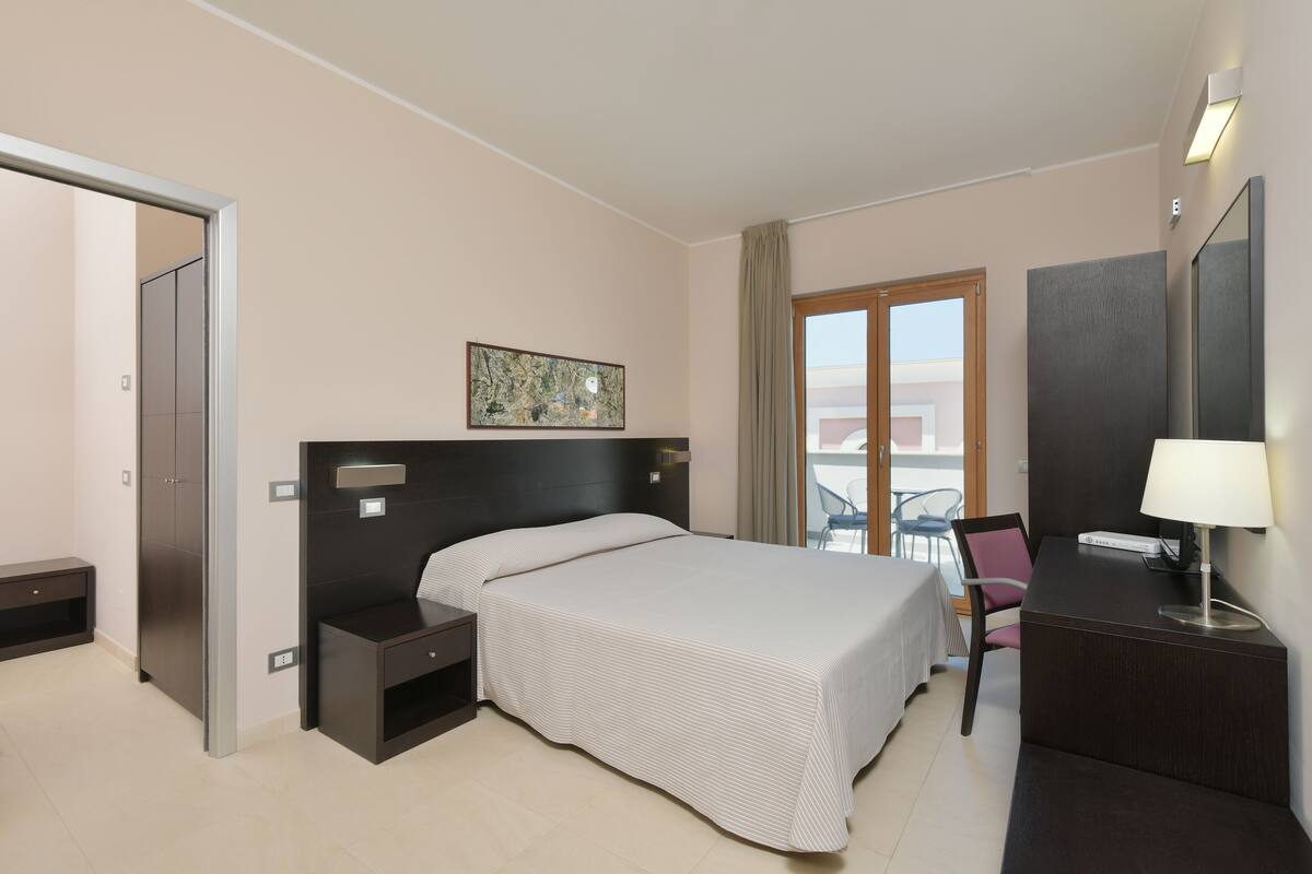 A hotel room with a bed, bedside tables, a desk with a lamp, a chair, and a large window with a view.