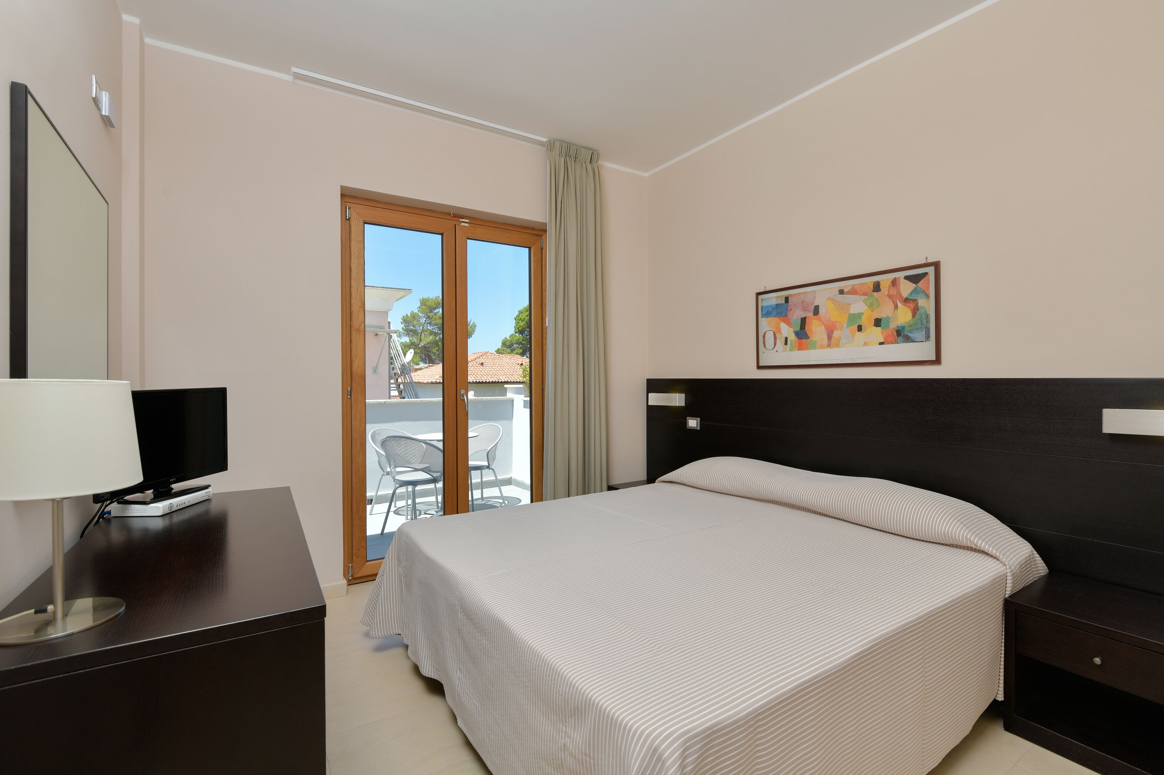 A hotel room with a bed, a TV on a desk, a painting on the wall, and a balcony with chairs.