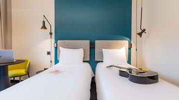 Quarto de hotel com duas camas, uma escrivaninha com um laptop, uma guitarra e uma cadeira amarela.