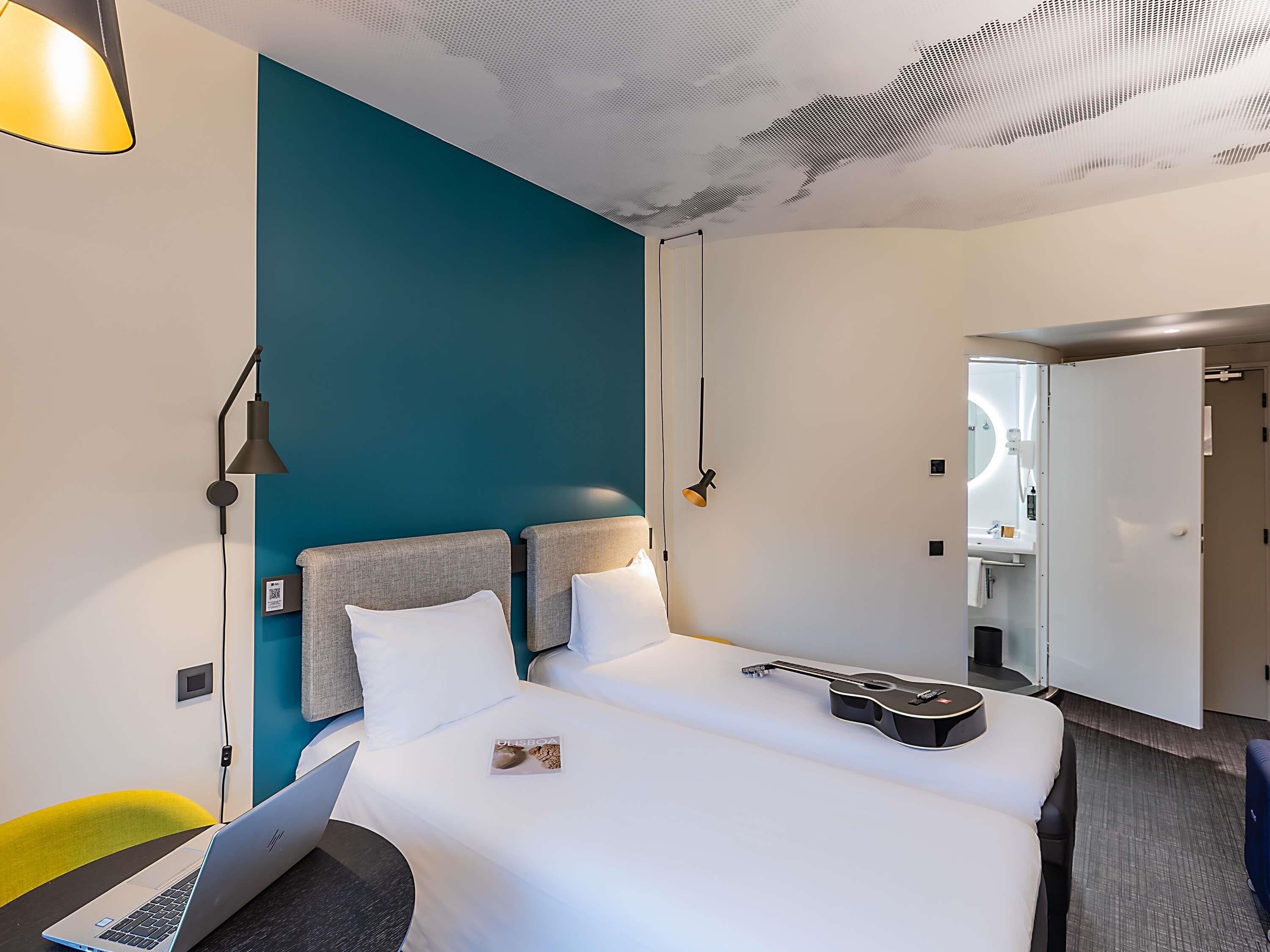 Una camera d'albergo con un letto, una scrivania con un portatile, una sedia, una lampada e una porta che dà su un bagno.