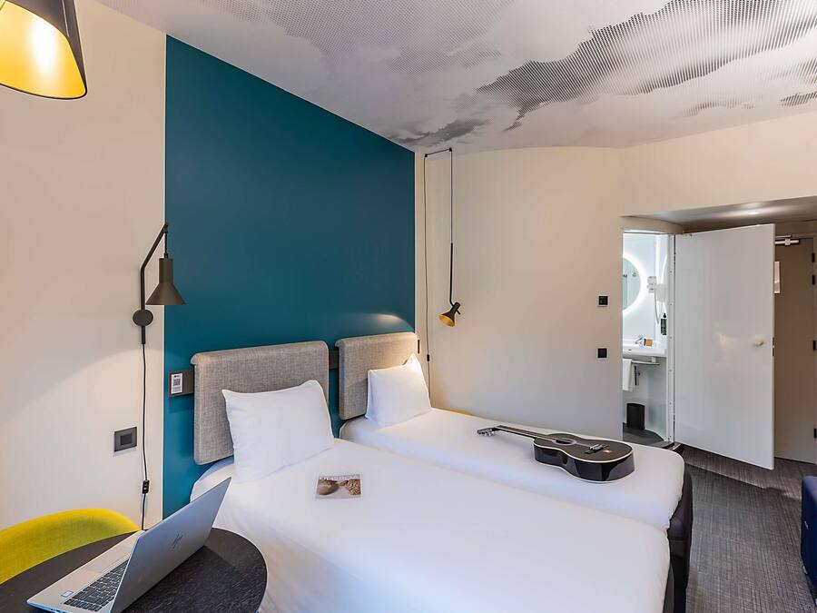 Una camera d'albergo con un letto, una scrivania con un portatile, una sedia, una lampada e una porta che dà su un bagno.
