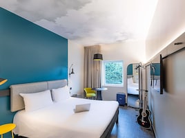 Habitación de hotel con cama, escritorio, silla, guitarra y ventana con vista a árboles.
