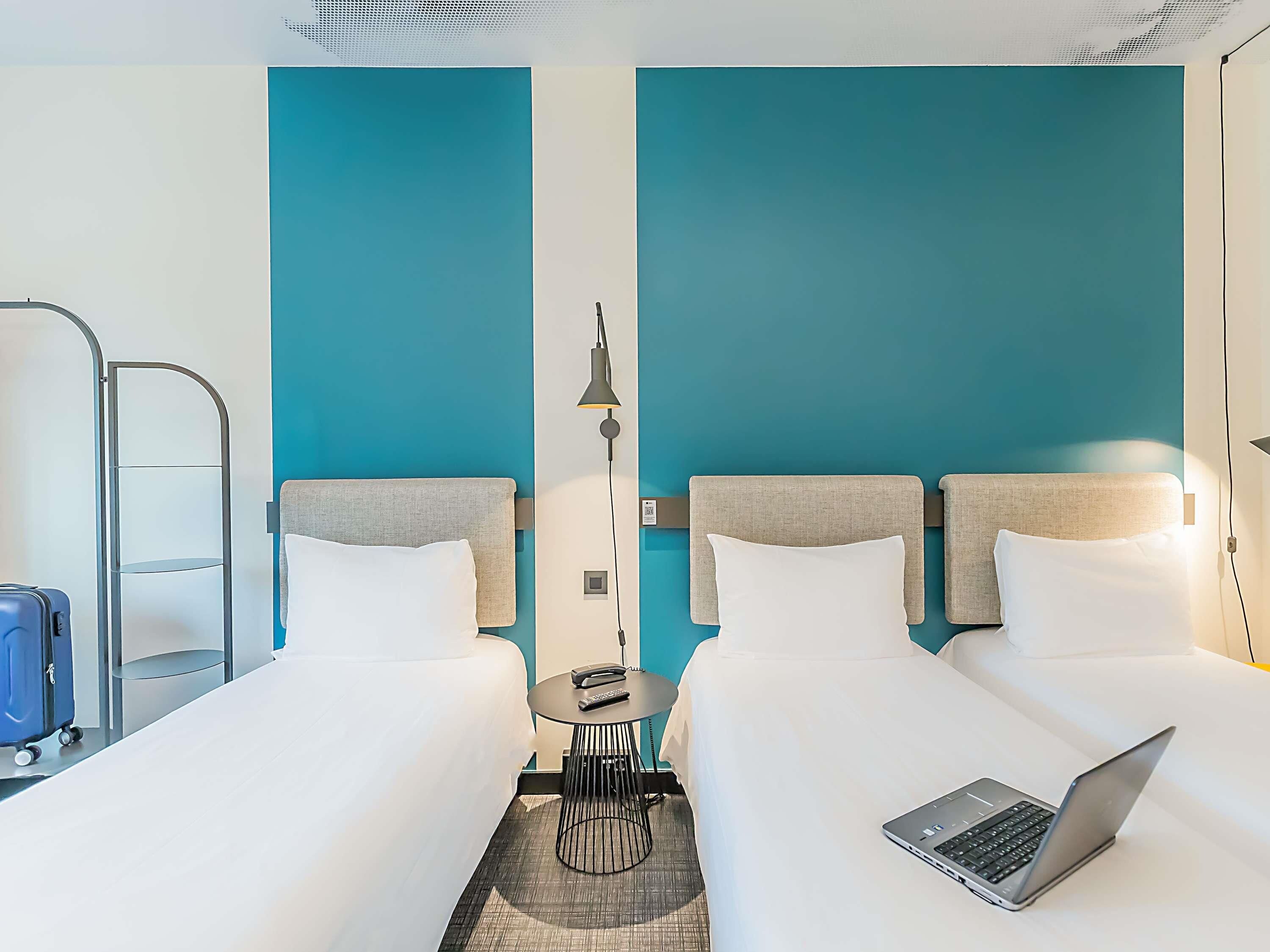 Ein Hotelzimmer mit zwei Betten, einem Schreibtisch, einem Laptop, einem Koffer und einer Lampe.