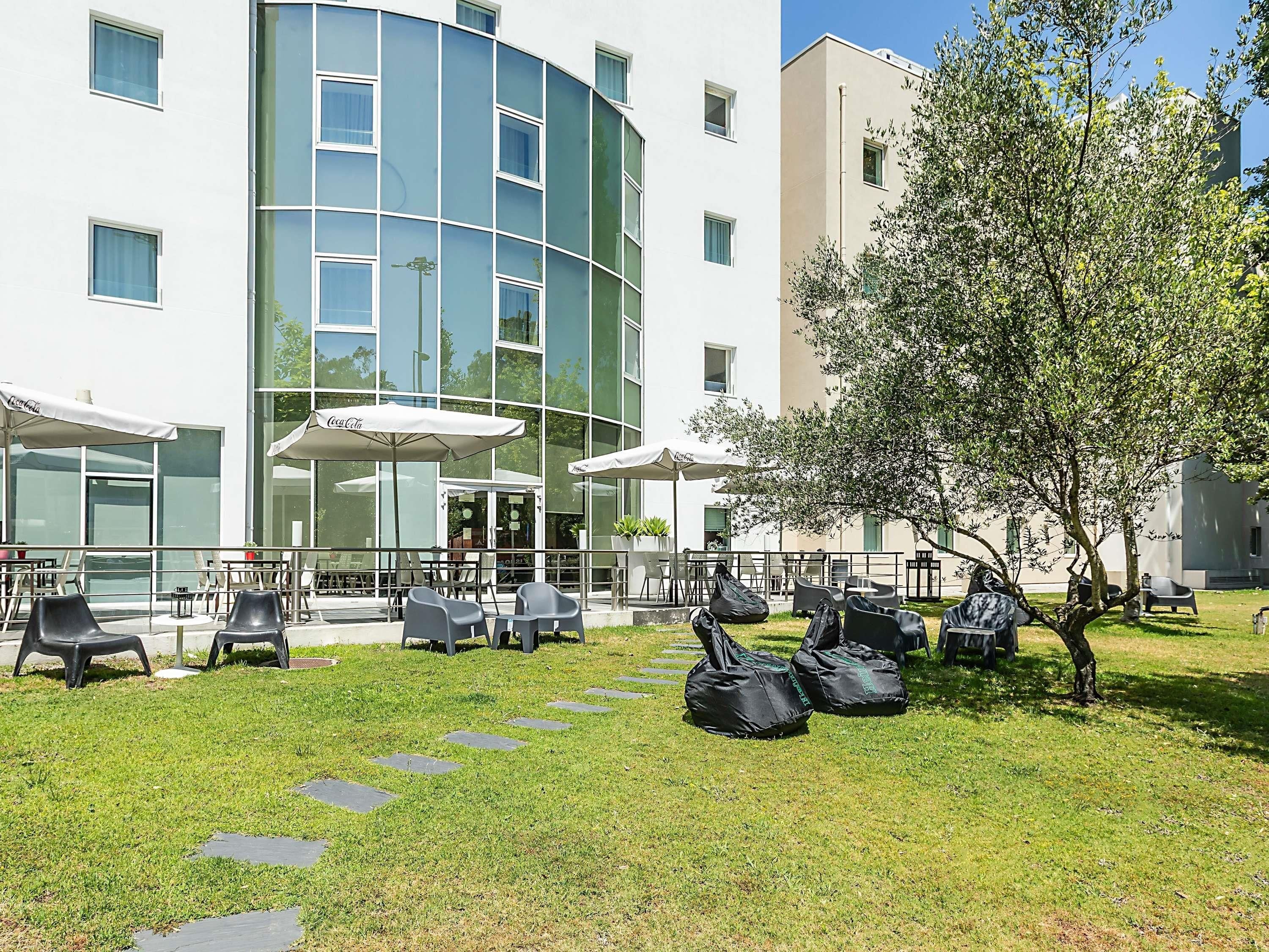 Un patio con mesas al aire libre, un paraguas con la marca Coca-Cola y un edificio con ventanas grandes y fachada curva.