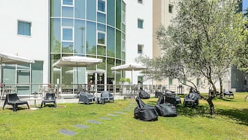 Un patio con mesas al aire libre, un paraguas con la marca Coca-Cola y un edificio con ventanas grandes y fachada curva.