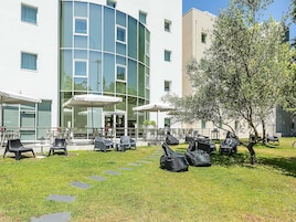Un patio con mesas al aire libre, un paraguas con la marca Coca-Cola y un edificio con ventanas grandes y fachada curva.