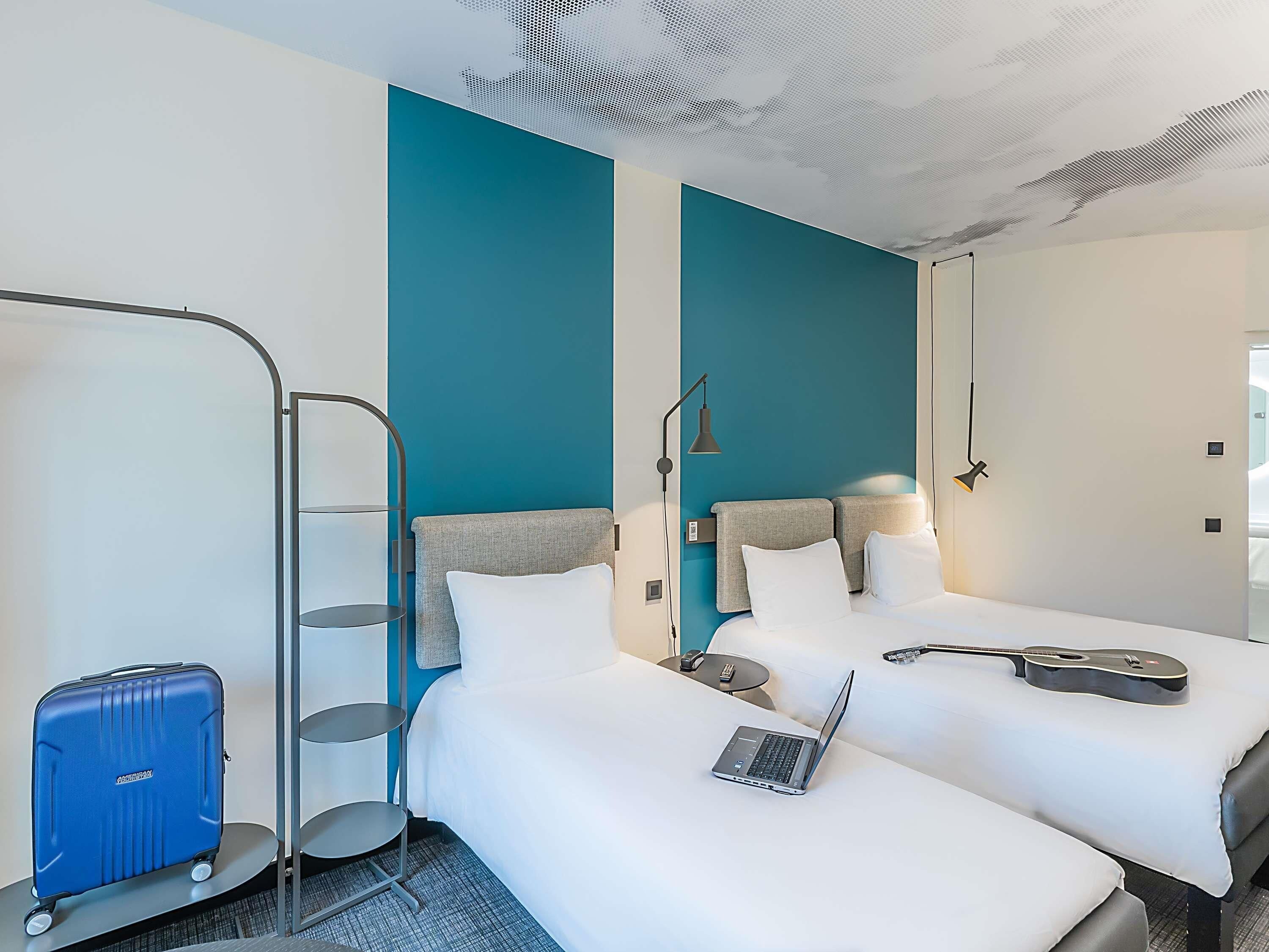 A hotel room with two beds, a laptop on one bed, a guitar on the other, and a suitcase on a trolley.