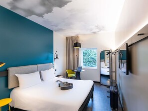 A modern hotel room with a large bed, a desk with a laptop, a mirror, and a suitcase. - ibis Lisboa Alfragide (Amadora)