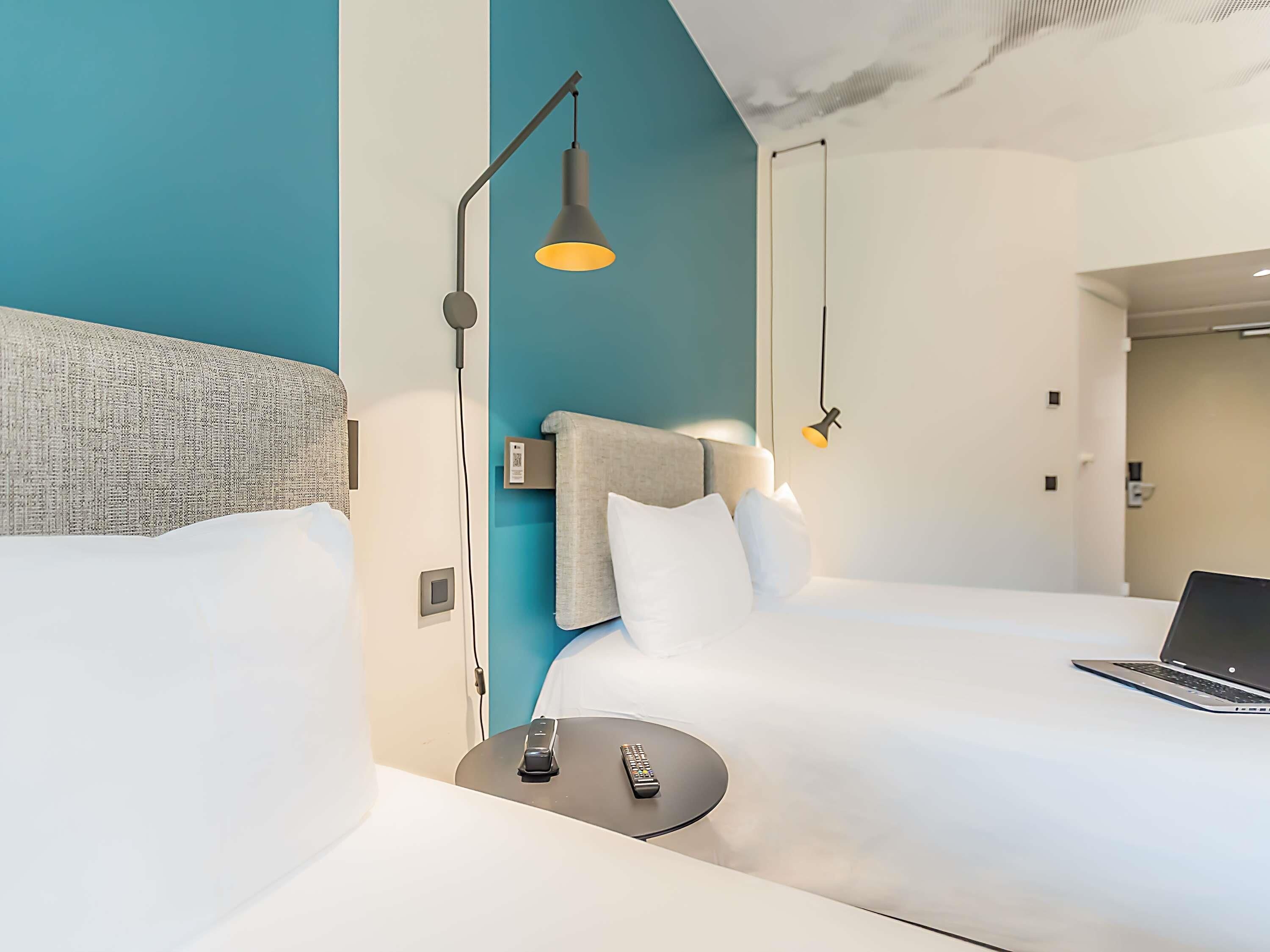 Ein modernes Hotelzimmer mit einem großen Bett, einem Schreibtisch mit einem Laptop, einem Nachttisch mit einer Fernbedienung und zwei Pendelleuchten.