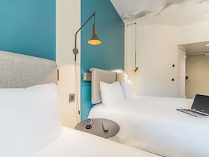 A modern hotel room with a large bed, a desk with a laptop, a bedside table with a remote control, and two pendant lights. - ibis Lisboa Alfragide (Amadora)