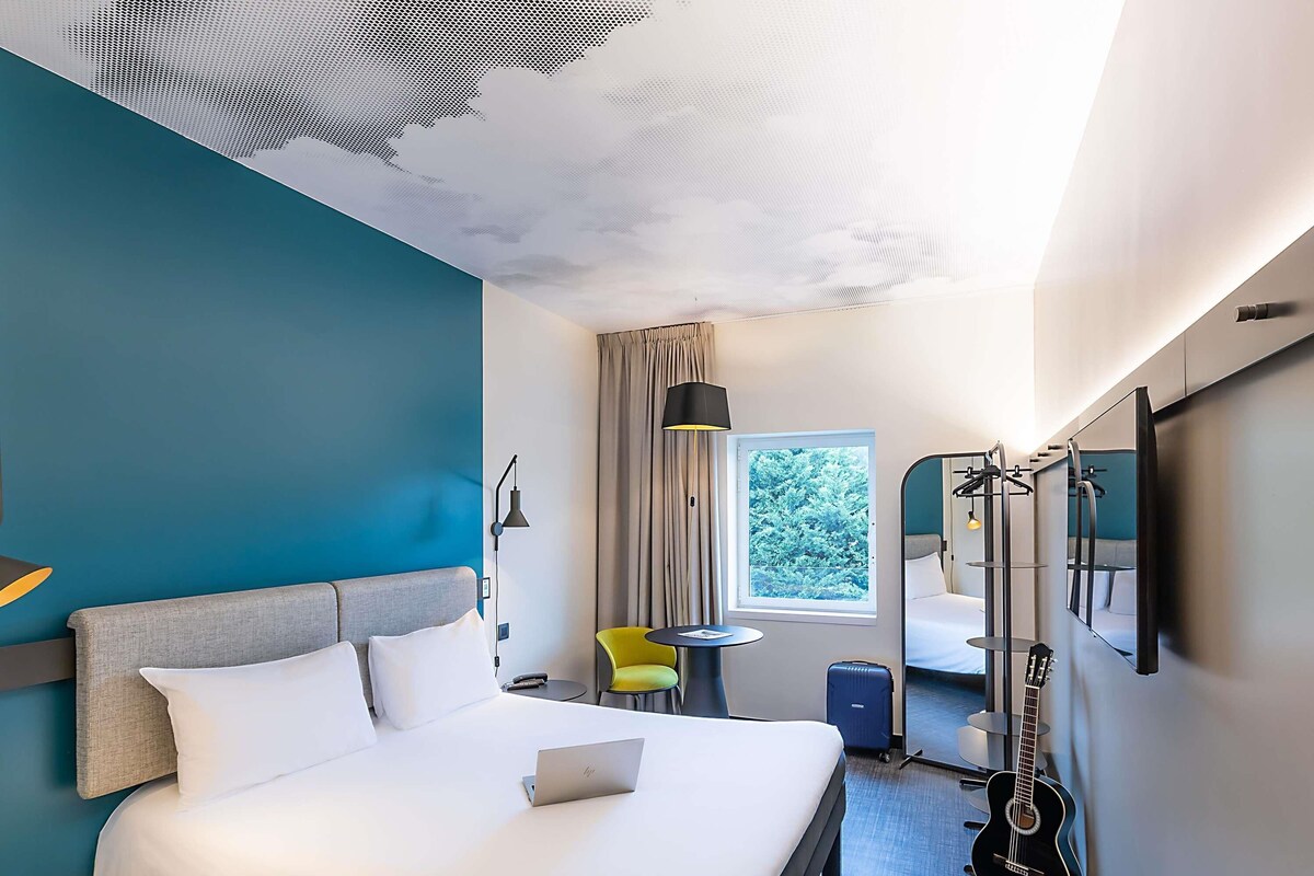 A hotel room with a bed, a desk, a chair, a guitar, a window with a view of trees, and a ceiling with a geometric pattern.