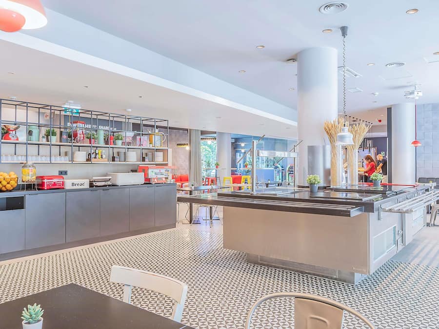 Un café moderno con una isla central, zona de estar y una cocina visible a través del muro de cristal.