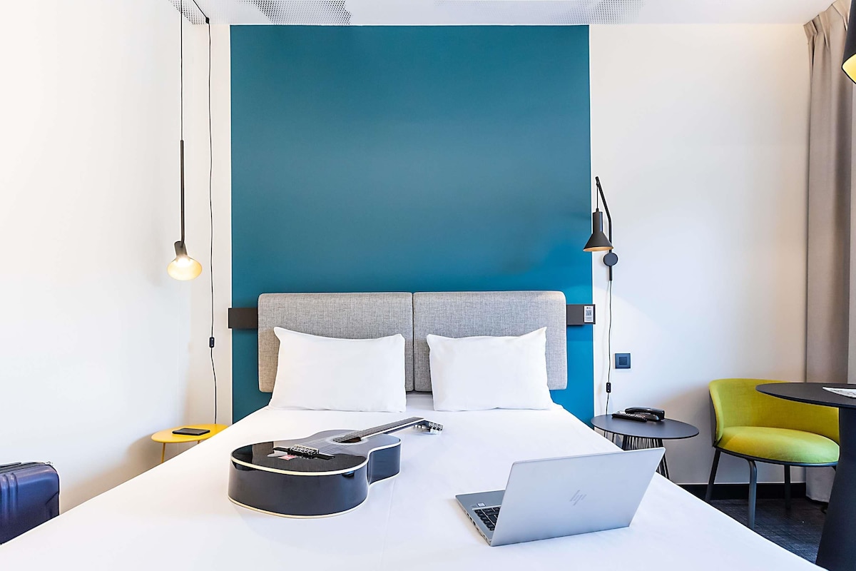 Una habitación de hotel con cama, ordenador portátil, guitarra, maleta y una silla.