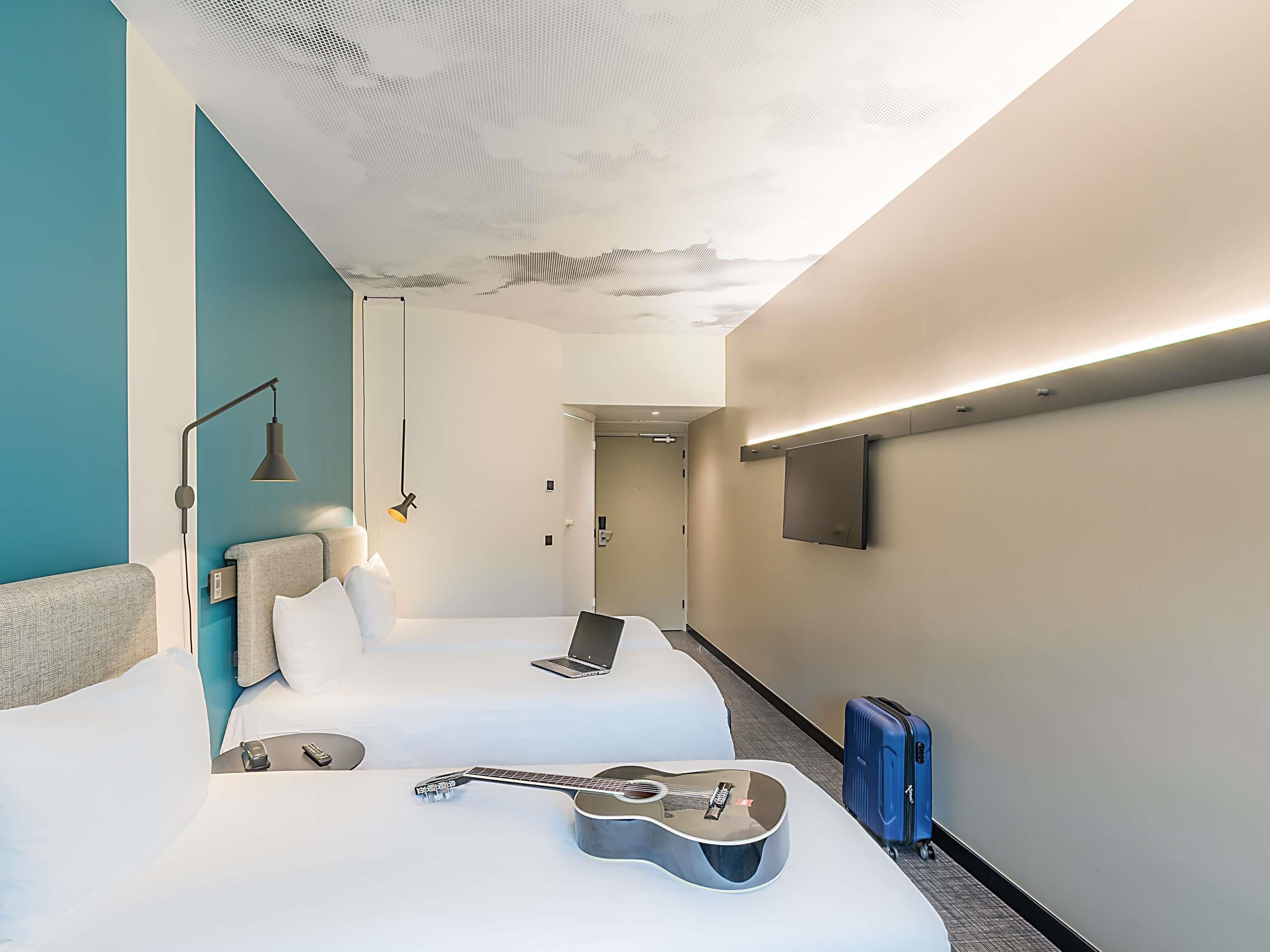 A hotel room with two beds, a guitar, a suitcase, a laptop, and a TV mounted on the wall.