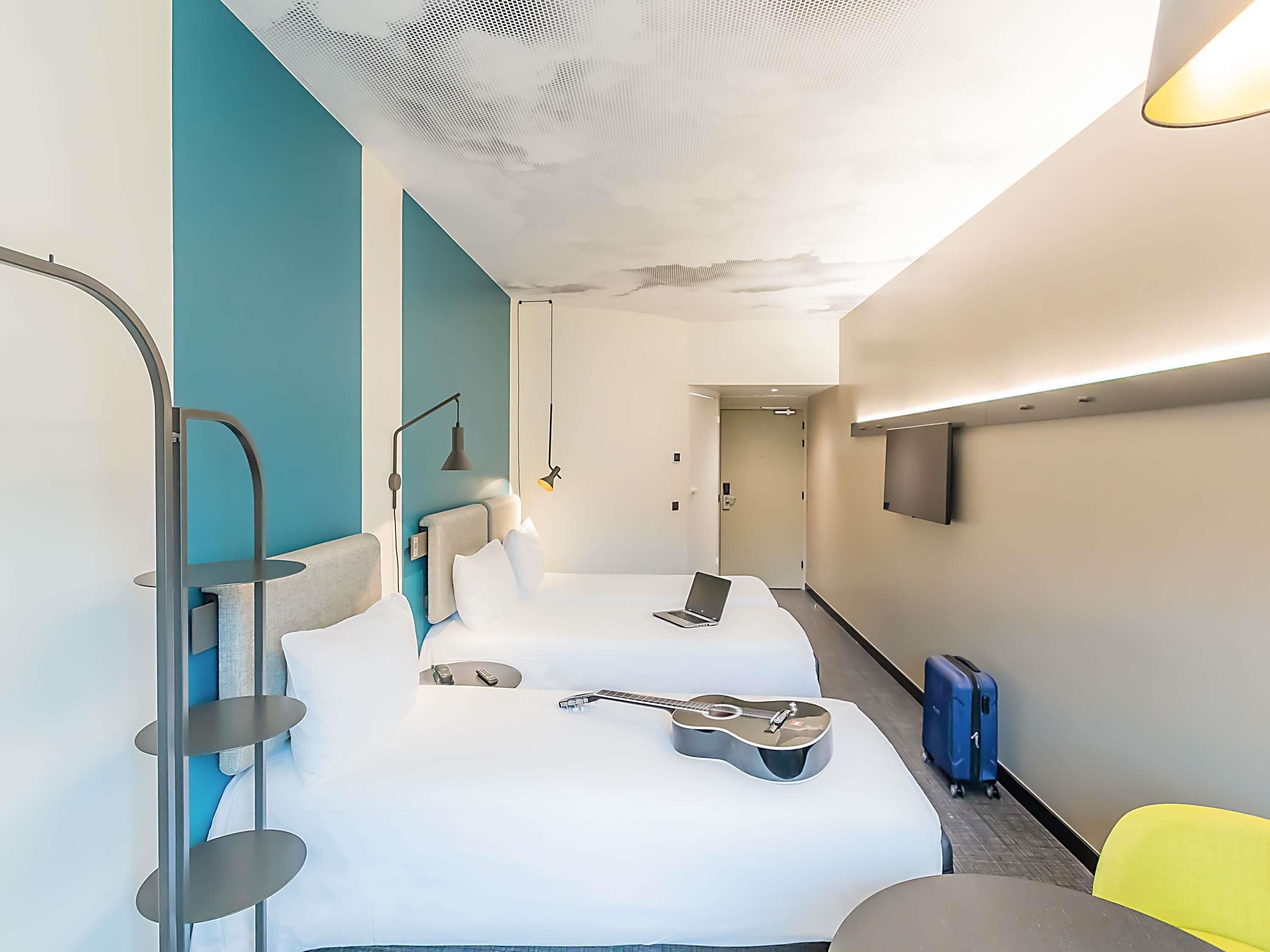 Quarto de hotel moderno com uma cama, um violão, um laptop e uma TV na parede.