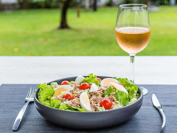 Un saladier rempli de salade composée, avec de la laitue, des tomates cerises, du thon et des œufs durs, accompagné d’un verre de vin sur une table.