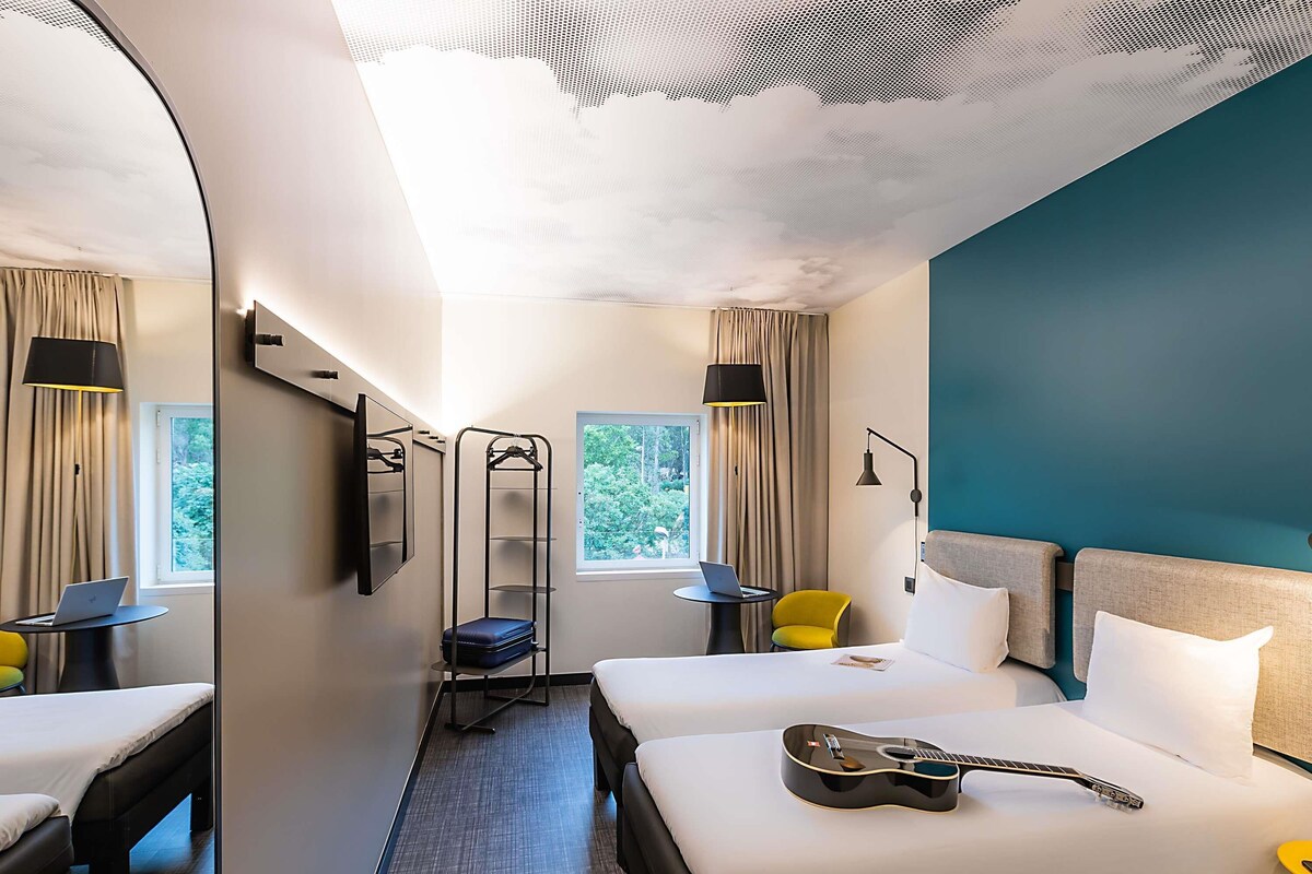 A hotel room with two beds, a guitar on one bed, a desk with a laptop, a chair, a hanging shelf, and a window with curtains.