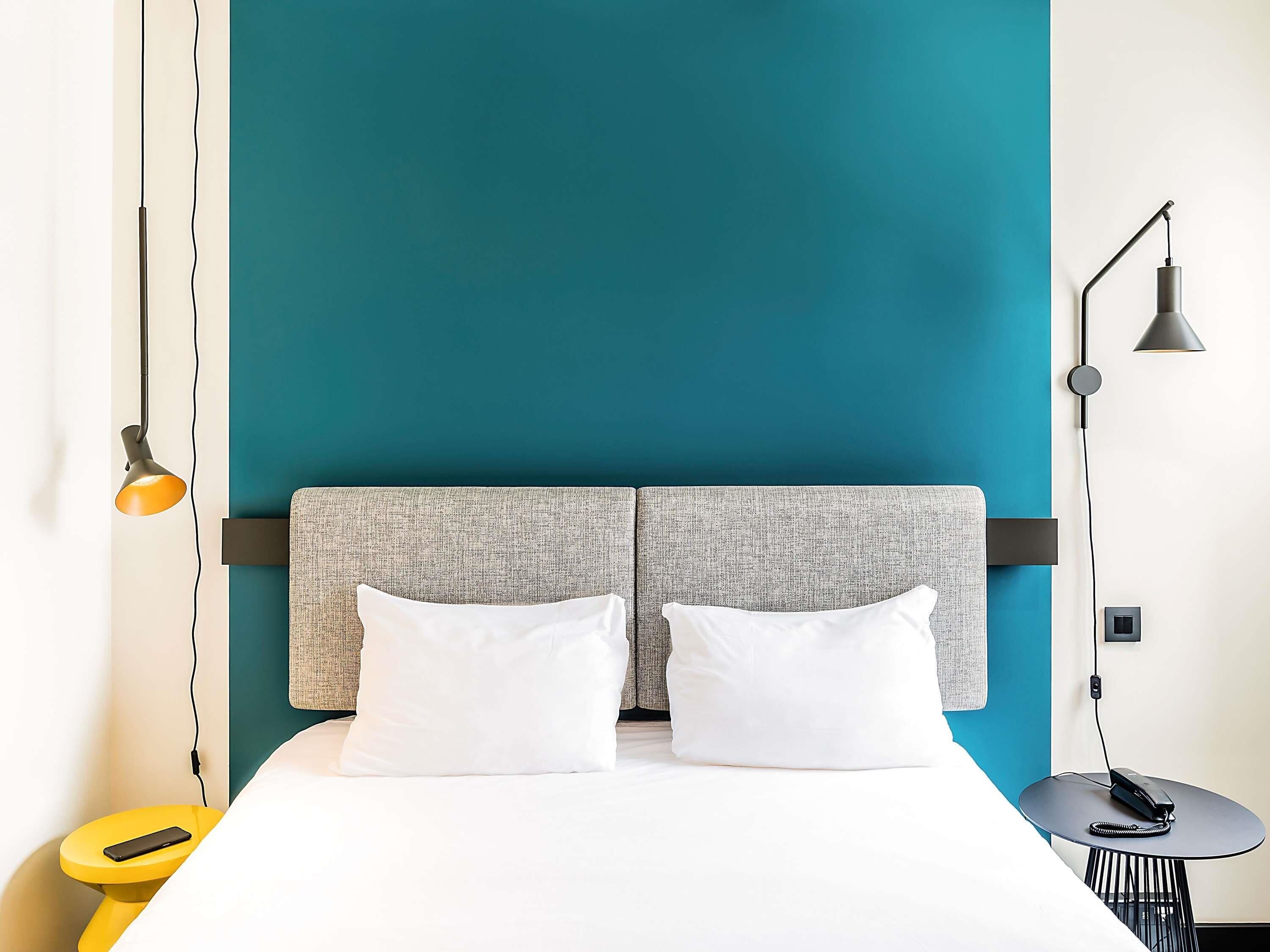 Et moderne hotelværelse med en teal farvet accentvæg, en grå tekstureret sengegavl, hvide sengetæpper, et gult natbord med en smartphone, og et lille rundt bord med en telefon.