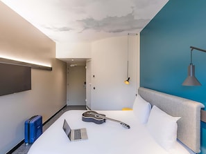 Una habitación de hotel moderna con una cama, una computadora portátil, una guitarra y una maleta azul.