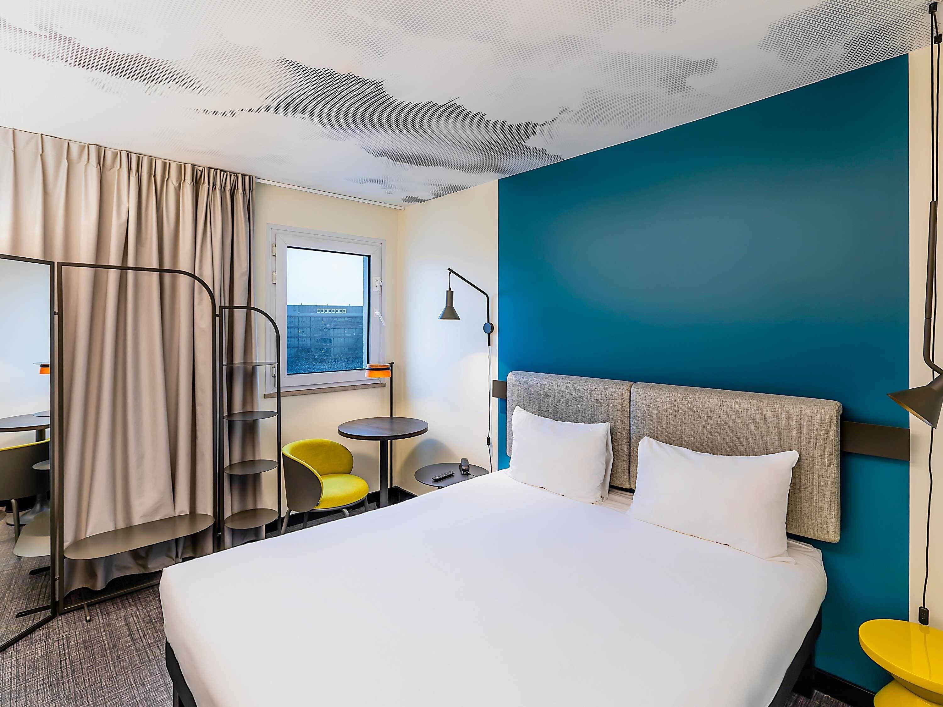 Quarto de hotel com cama, escrivaninha, cadeira, mesa pequena e janela.