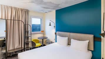 Quarto de hotel com cama, escrivaninha, cadeira, mesa pequena e janela.
