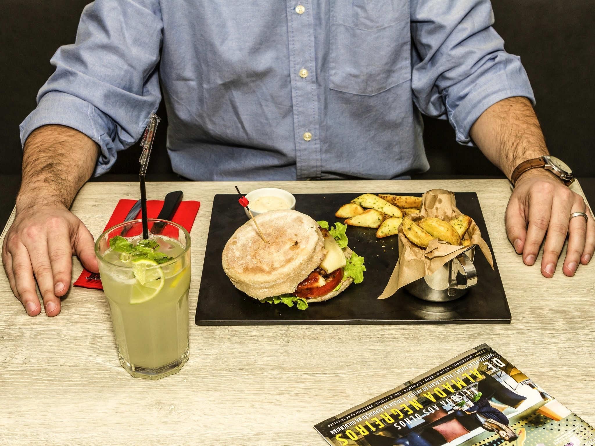 Een persoon zit aan een tafel met een maaltijd bestaande uit een sandwich, frietjes en een drankje, met een menukaart en een tijdschrift op de tafel.