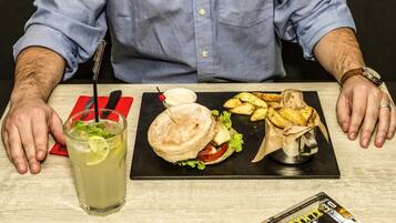 Een persoon zit aan een tafel met een maaltijd bestaande uit een sandwich, frietjes en een drankje, met een menukaart en een tijdschrift op de tafel.