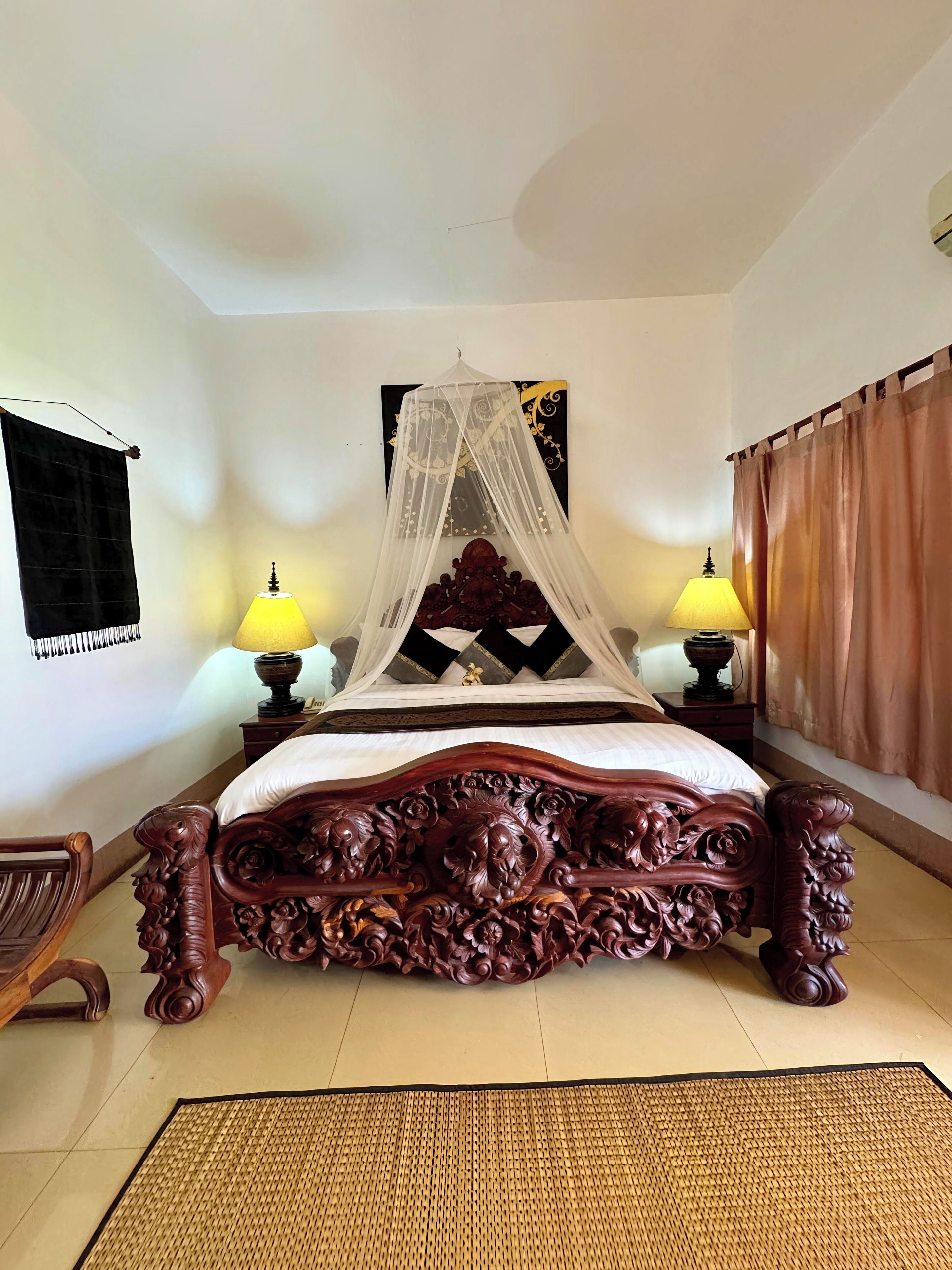 A four-poster bed with a canopy, two bedside lamps, a decorative headboard, a woven mat, and a window with a curtain.