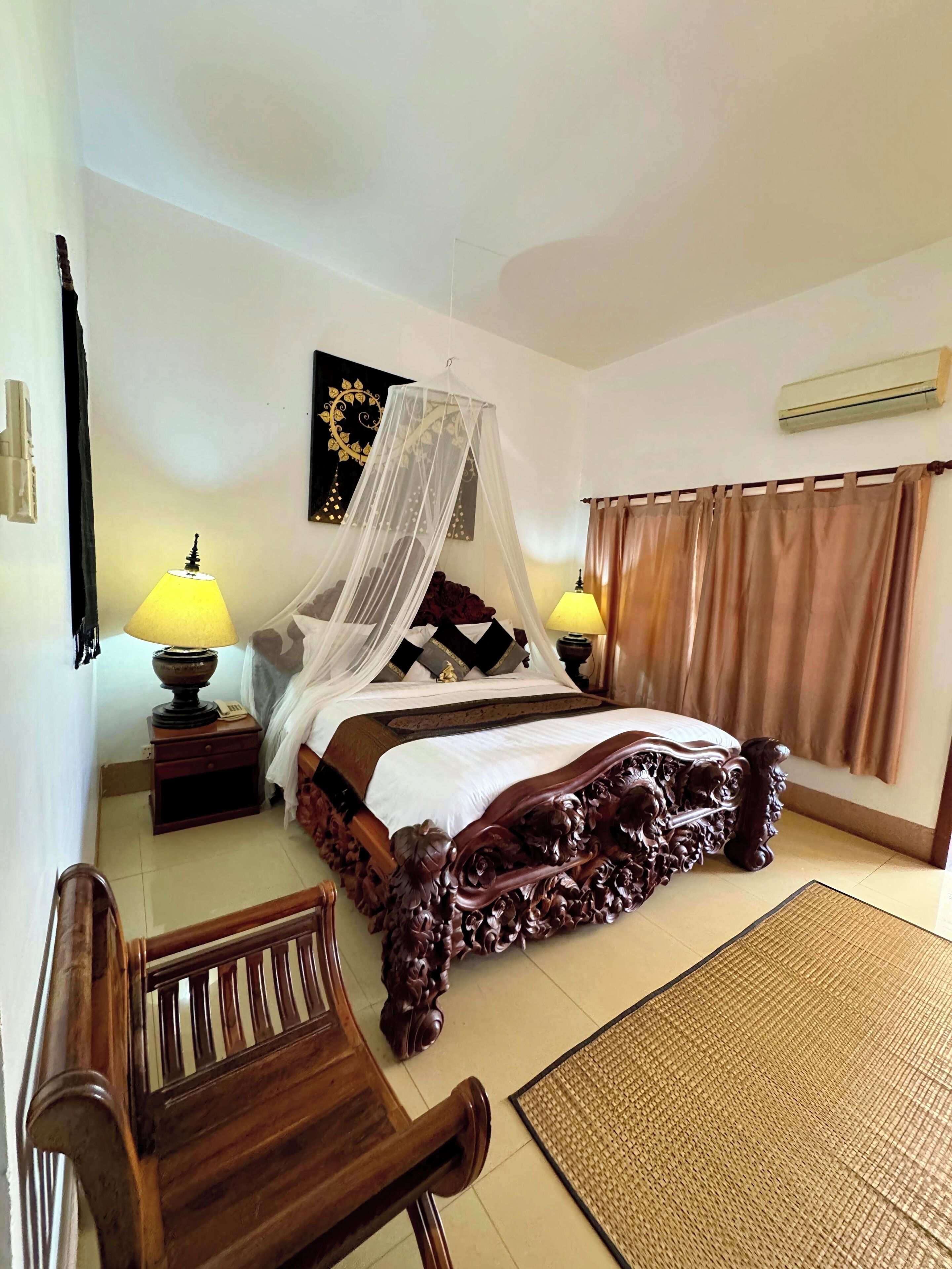 A bedroom with a four-poster bed, two bedside lamps, a decorative wall hanging, and a large window with curtains.