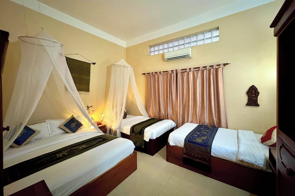 A hotel room with two single beds, a mosquito net over one bed, a ceiling fan, a window with curtains, and an air conditioning unit.
