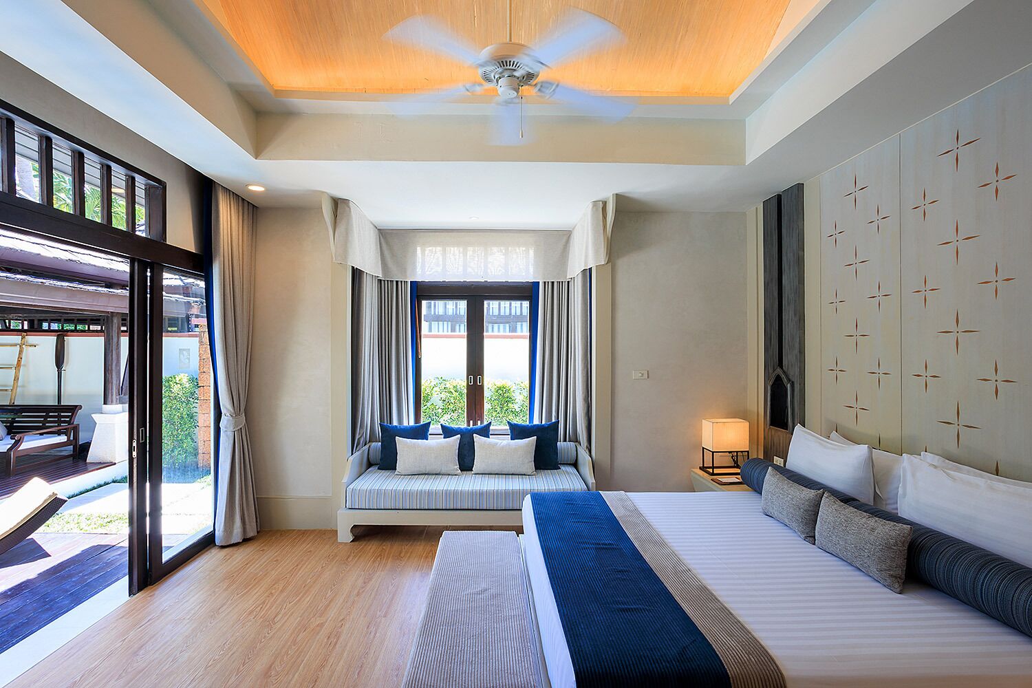 Habitación de hotel con una cama grande, sofá cama, ventilador de techo, ventana con cortinas y vistas al exterior.