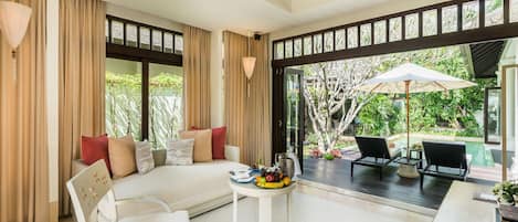 A spacious room with a large window, a sofa, a dining table with fruit, and a view of a pool and greenery.