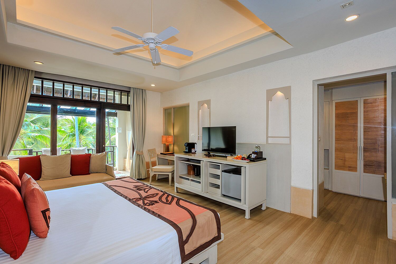 A hotel room with a bed, a desk with a television, a ceiling fan, and a view of greenery outside.