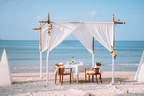 Une cérémonie de mariage organisée sur la plage, avec une tente blanche, des chaises en bois et une table recouverte d’une nappe blanche.
