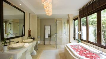 A modern bathroom with two sinks, a large bathtub with red flower petals, a glass shower enclosure, and a view of greenery outside.