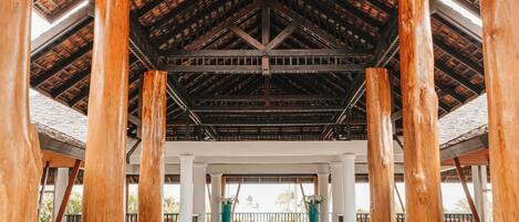 A spacious hall with high wooden beams and columns, a patterned floor, and a view of the outside.
