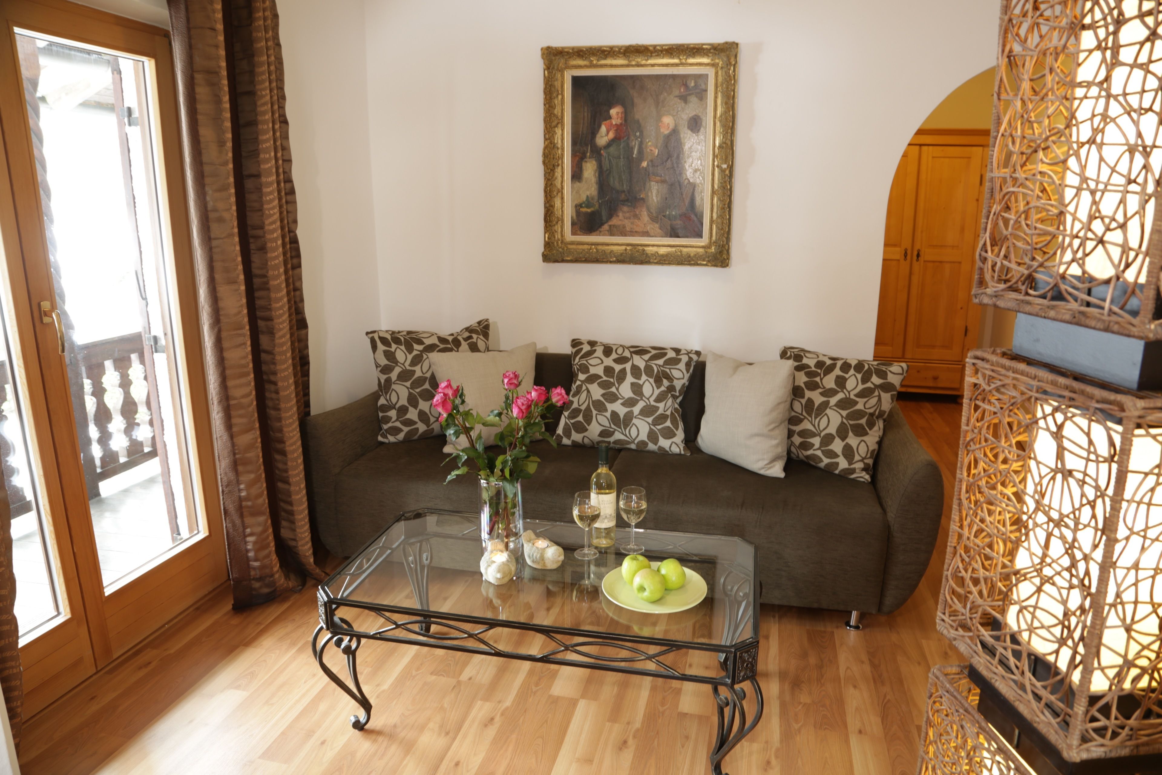 A living room with a glass coffee table, a sofa with patterned cushions, a painting on the wall, and a wicker lamp.