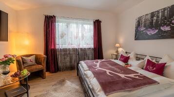 Un dormitorio con cama, una silla, una mesa con un florero y un cuadro en la pared.