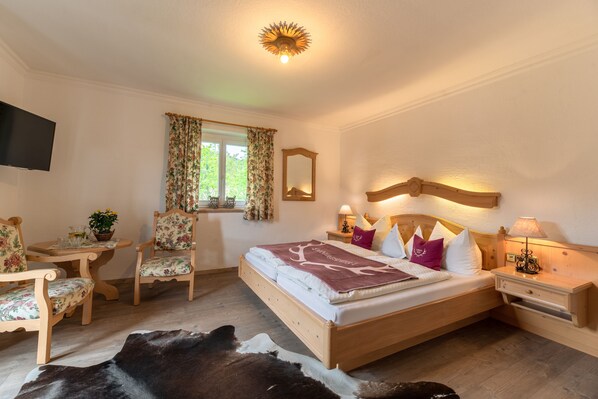 A bedroom with a wooden bed, floral chairs, a small table, a TV, and a window with curtains.