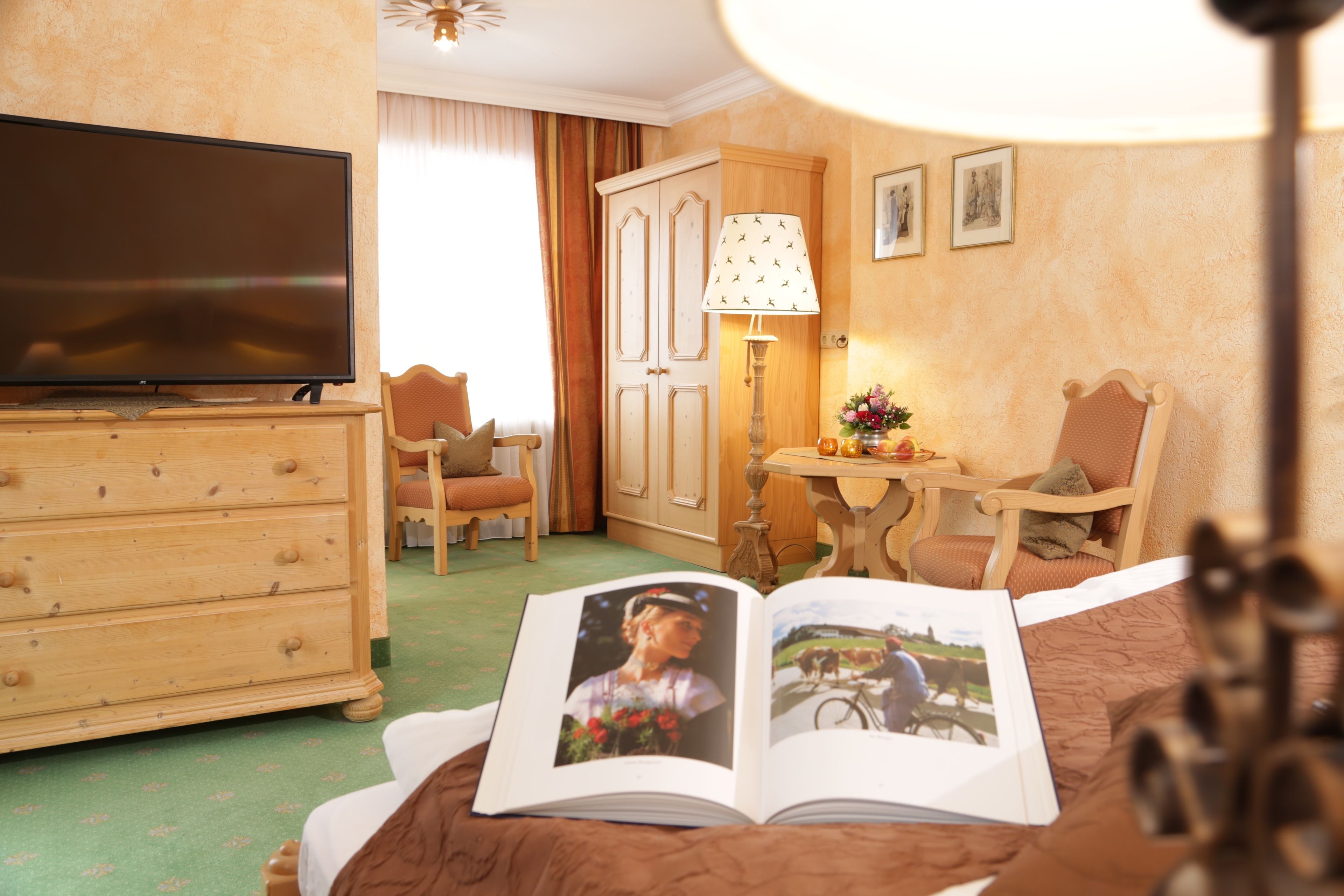 Une chambre d’hôtel équipée d’une télévision, d’une commode en bois, de deux chaises, d’une table avec une lampe et d’un livre posé sur le lit.